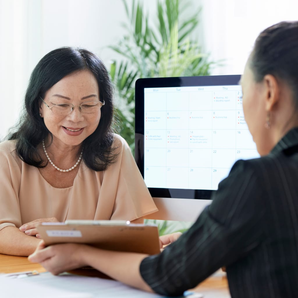 Two women in a professional setting discussing documents, with one holding a clipboard and the other smiling, in front of a computer displaying a calendar, emphasizing collaboration and communication in property management.
