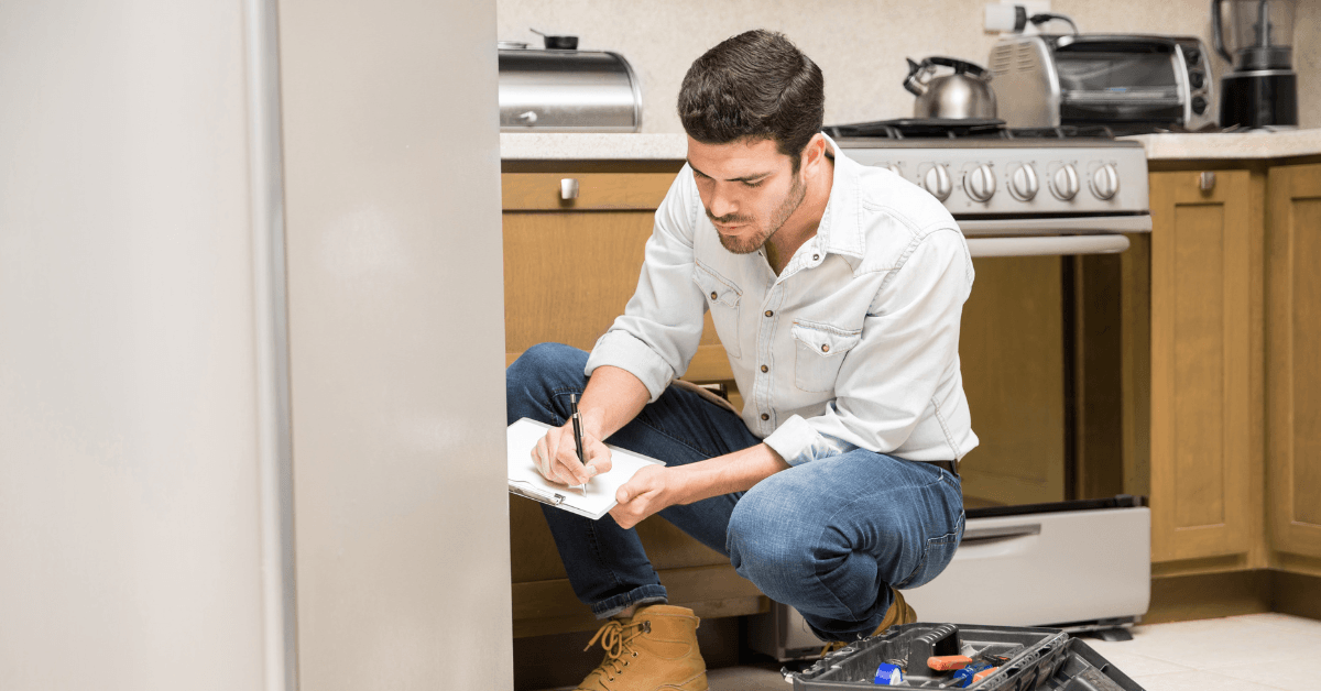 Man crouching in kitchen, taking notes while inspecting appliances, highlighting proactive maintenance in property management.