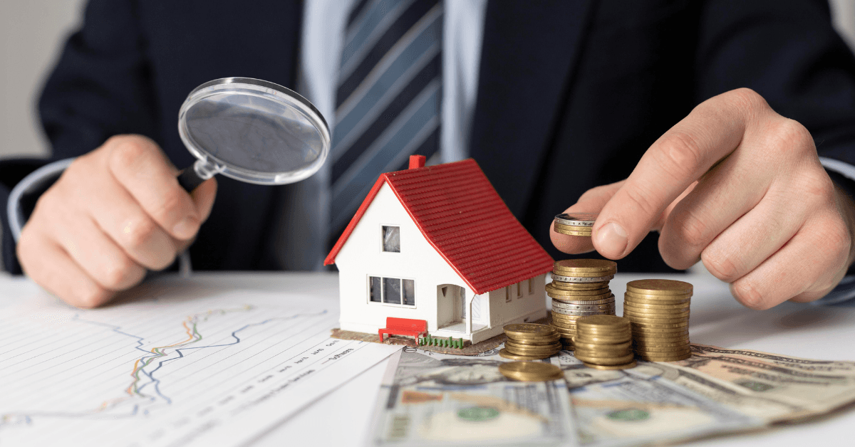 Businessman analyzing property investment with magnifying glass, model house, and stacks of coins and cash, illustrating financial management in real estate.