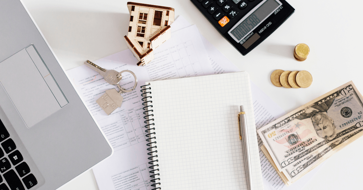 Laptop, house model, keys, notepad, calculator, coins, and cash on paperwork, symbolizing property management and investment strategies.