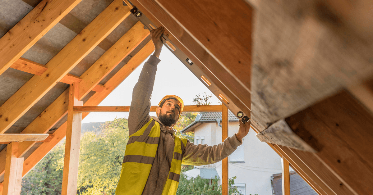 Construction worker measuring roof structure for dormer addition in multifamily home renovation, emphasizing property management and value enhancement.