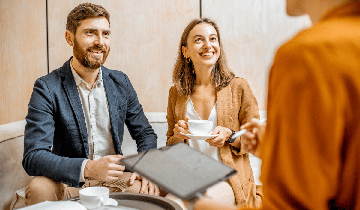 A man and a woman are sitting on a couch, having coffee with another person standing in front of them.