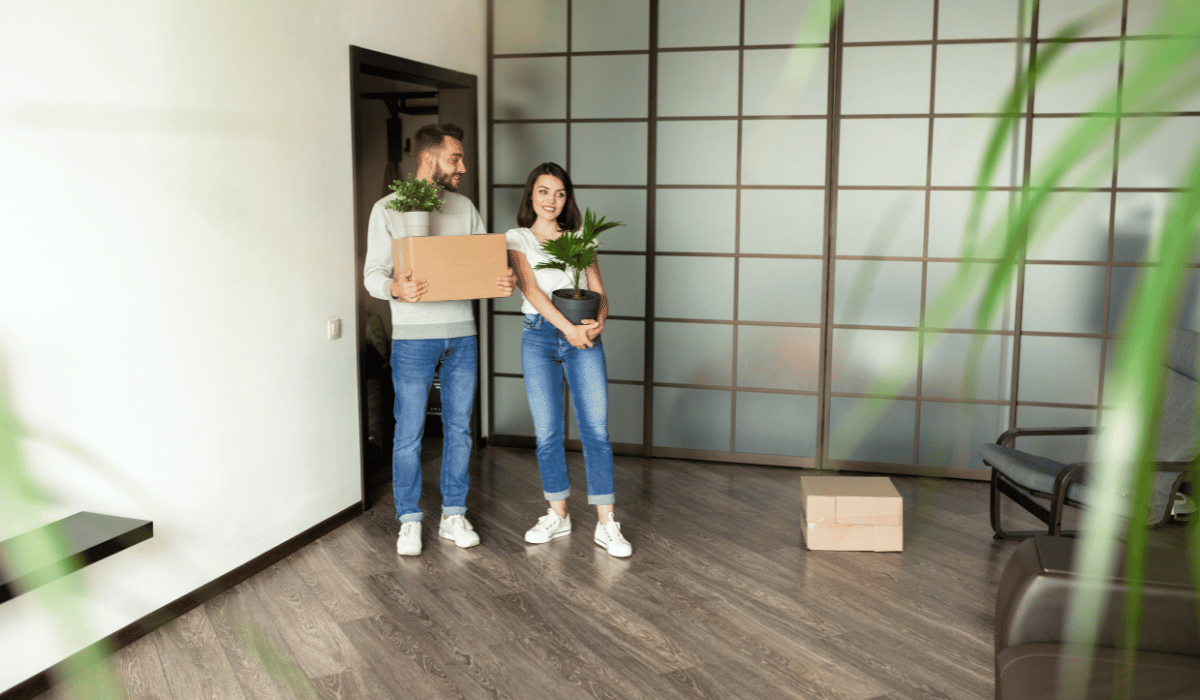 A couple is moving, holding boxes and plants, with a box on the floor.