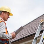 A worker on a ladder checks roof shingles while holding a clipboard