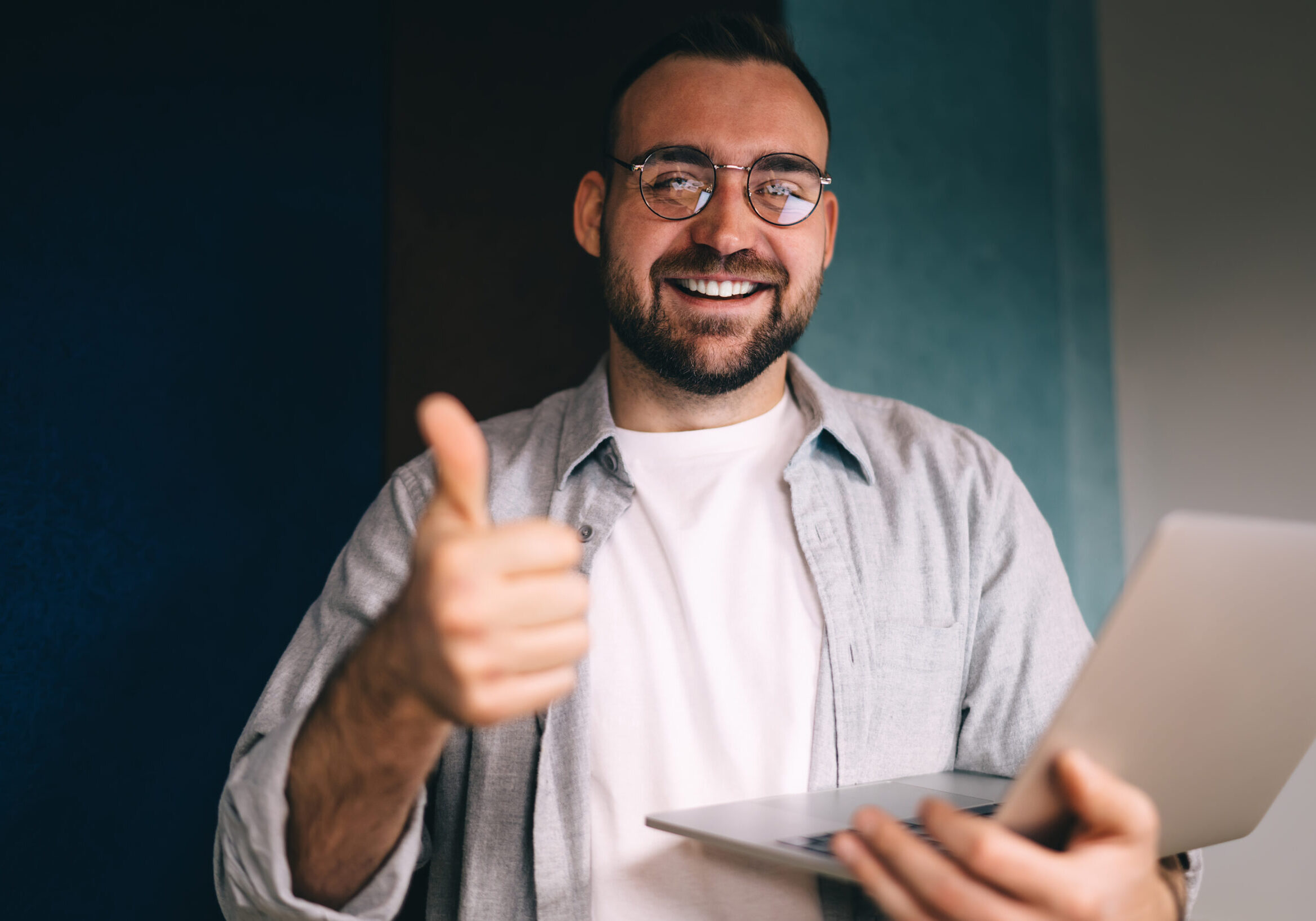 Positive male freelancer in casual outfit and glasses holding netbook and showing like gesture looking at camera standing against striped wall